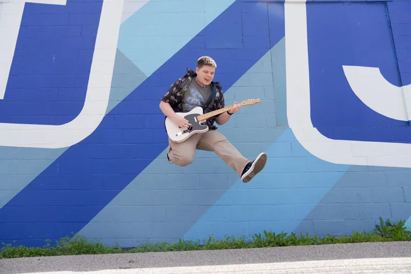 A young jumps in the air in front of a blue wall holding a guitar