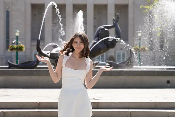 A young woman in a white dress standing with her arms up in front of a sculpture fountain