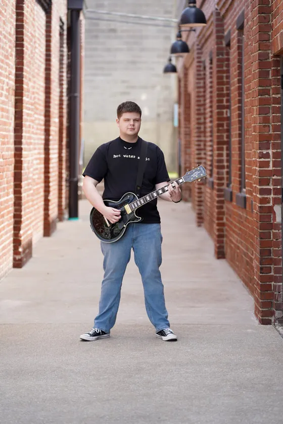 A young man stands in an alley holding a guitar